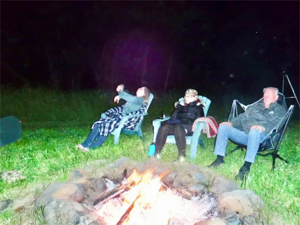 Three people sitting around a glowing campfire at night, gazing upward under a dark sky filled with subtle orb-like lights.