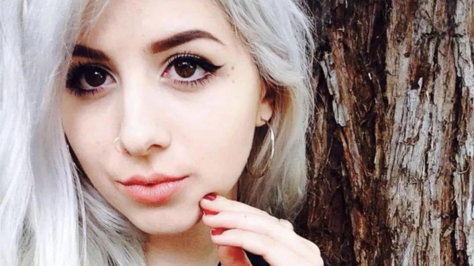 Brooke, a young woman with platinum blonde hair and winged eyeliner, gently resting her hand on her chin in front of a textured tree trunk.