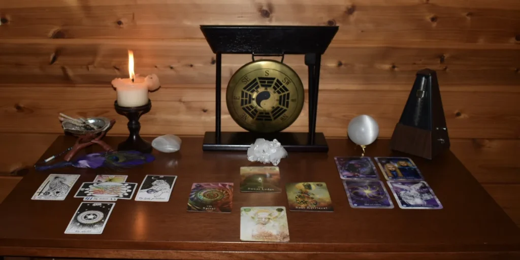 A spiritual reading setup on a wooden table featuring Tarot and Oracle cards laid out in three distinct spreads. A lit candle, crystal cluster, sage bundle with feathers, crystal spheres, a metronome, and a decorative gong with yin-yang and directional symbols create a sacred and intentional atmosphere.
